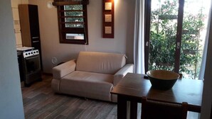 A living room with a sofa, a dining table with a bowl, a stove, and a window with blinds. - Vila Tamu (Imbituba)