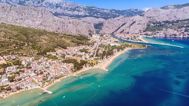 Vista aérea de un pueblo costero con playa, edificios residenciales y terreno montañoso al fondo.