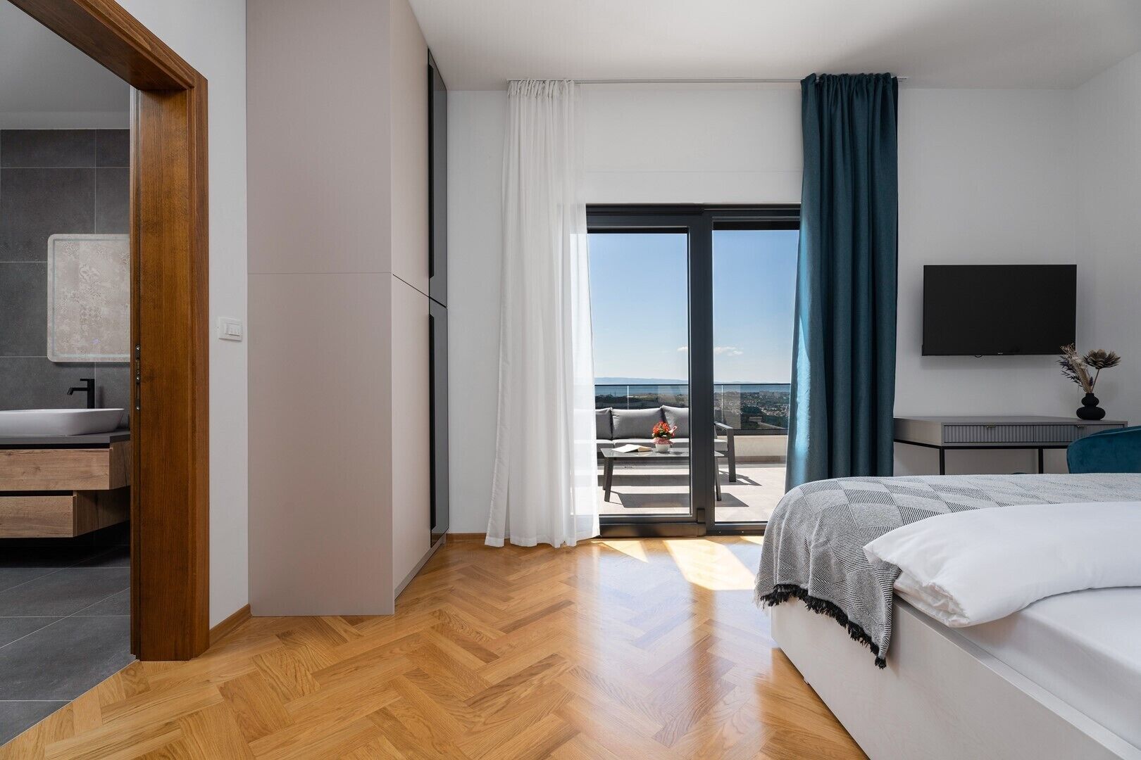A modern hotel room with a wooden floor, a bed, a TV, a balcony with a view, and a bathroom with a sink and mirror.