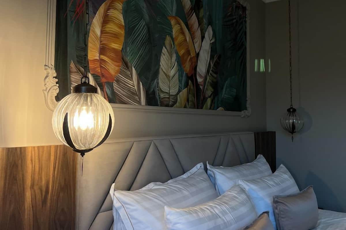 A bedroom with a bed, pillows, a wall-mounted light fixture, and a painting of feathers.