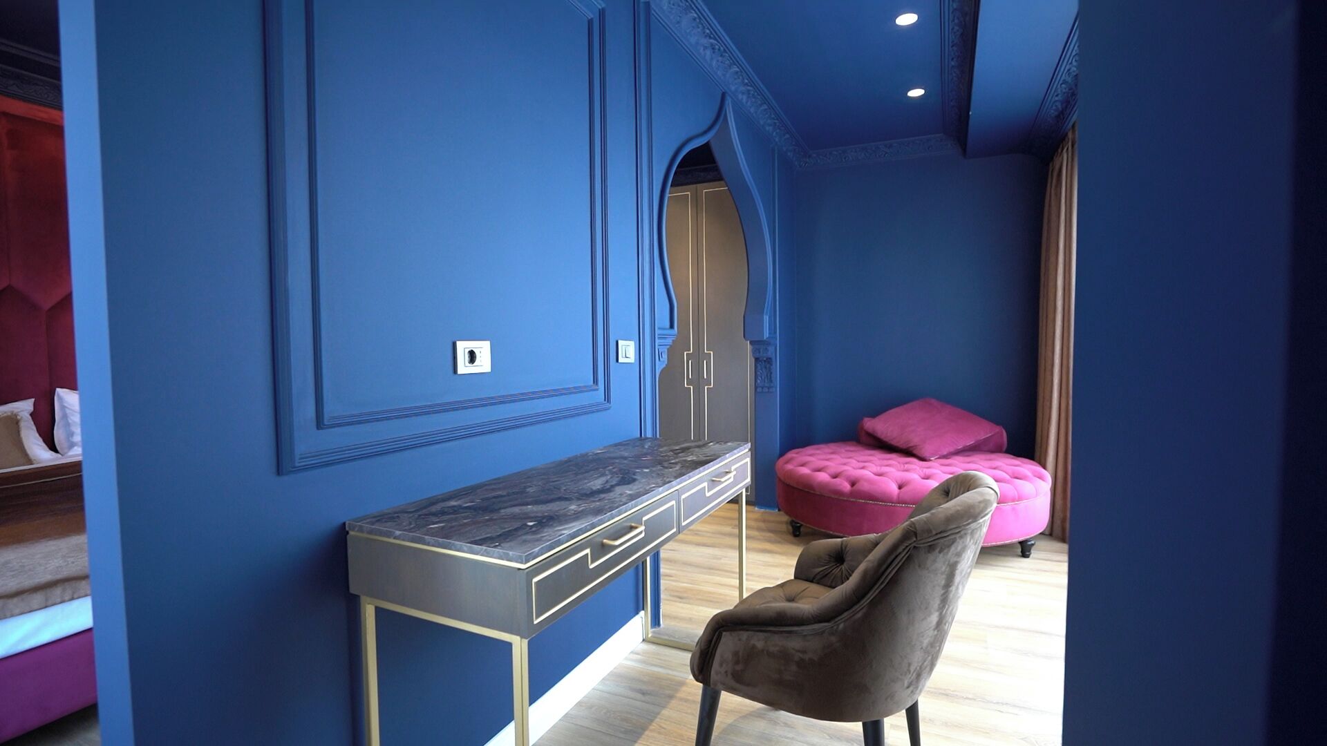 Una habitación con pared azul, un escritorio de mármol, un otomano rosa y una silla gris.