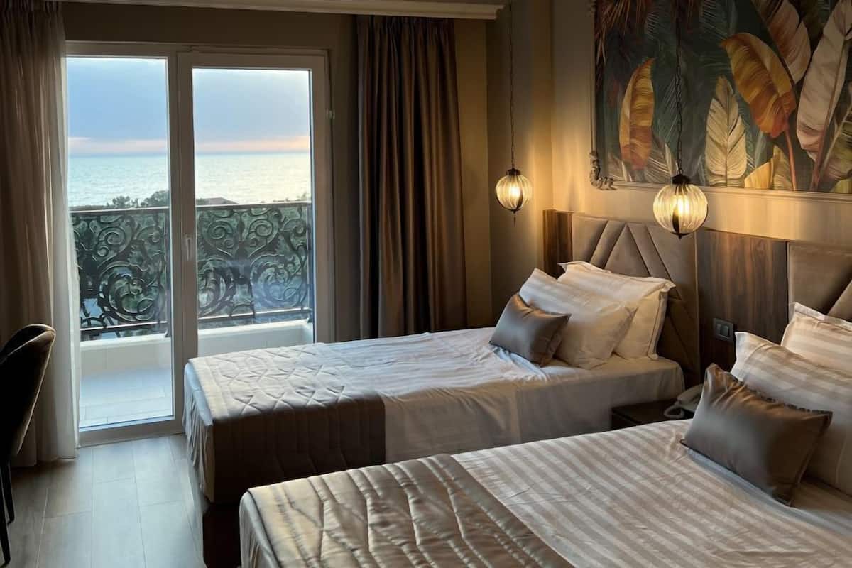 A hotel room with two beds, a view of the sea through a balcony, and a painting on the wall.