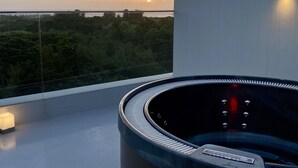 A modern bathroom with a circular tub, a glass-enclosed balcony, and a view of the sunset over the sea. - SkyBlueVilla (読谷村)