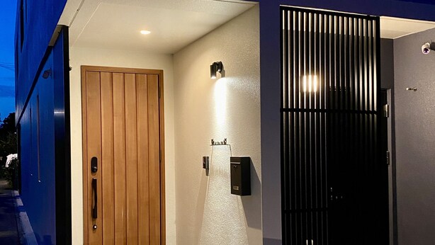 A modern building entrance with a wooden door, a black gate, and a light fixture on the wall. - SkyBlueVilla (読谷村)