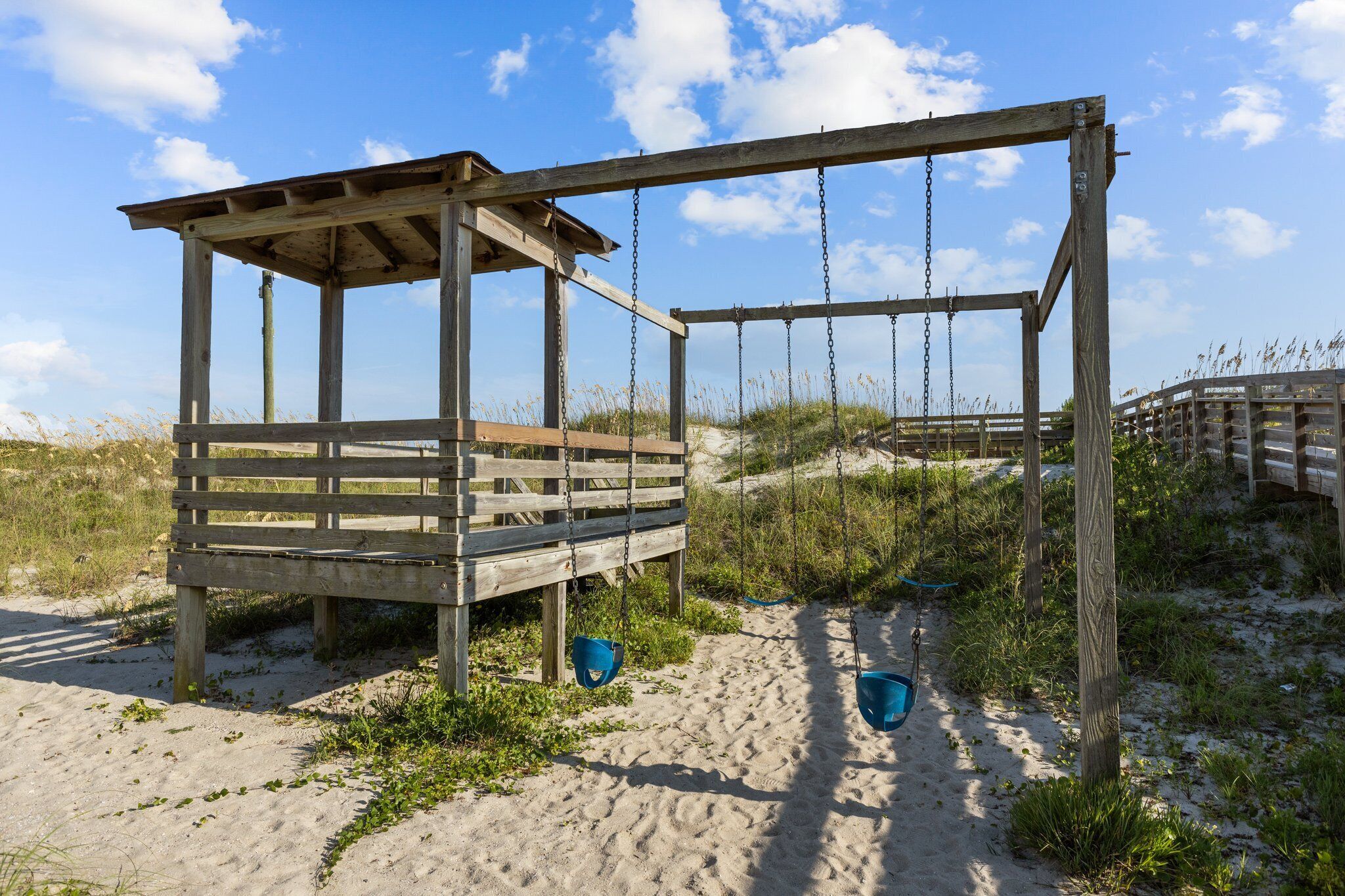 Une balançoire en bois installée sur une plage, avec un banc, et située sur un terrain sablonneux, avec des dunes en arrière-plan.