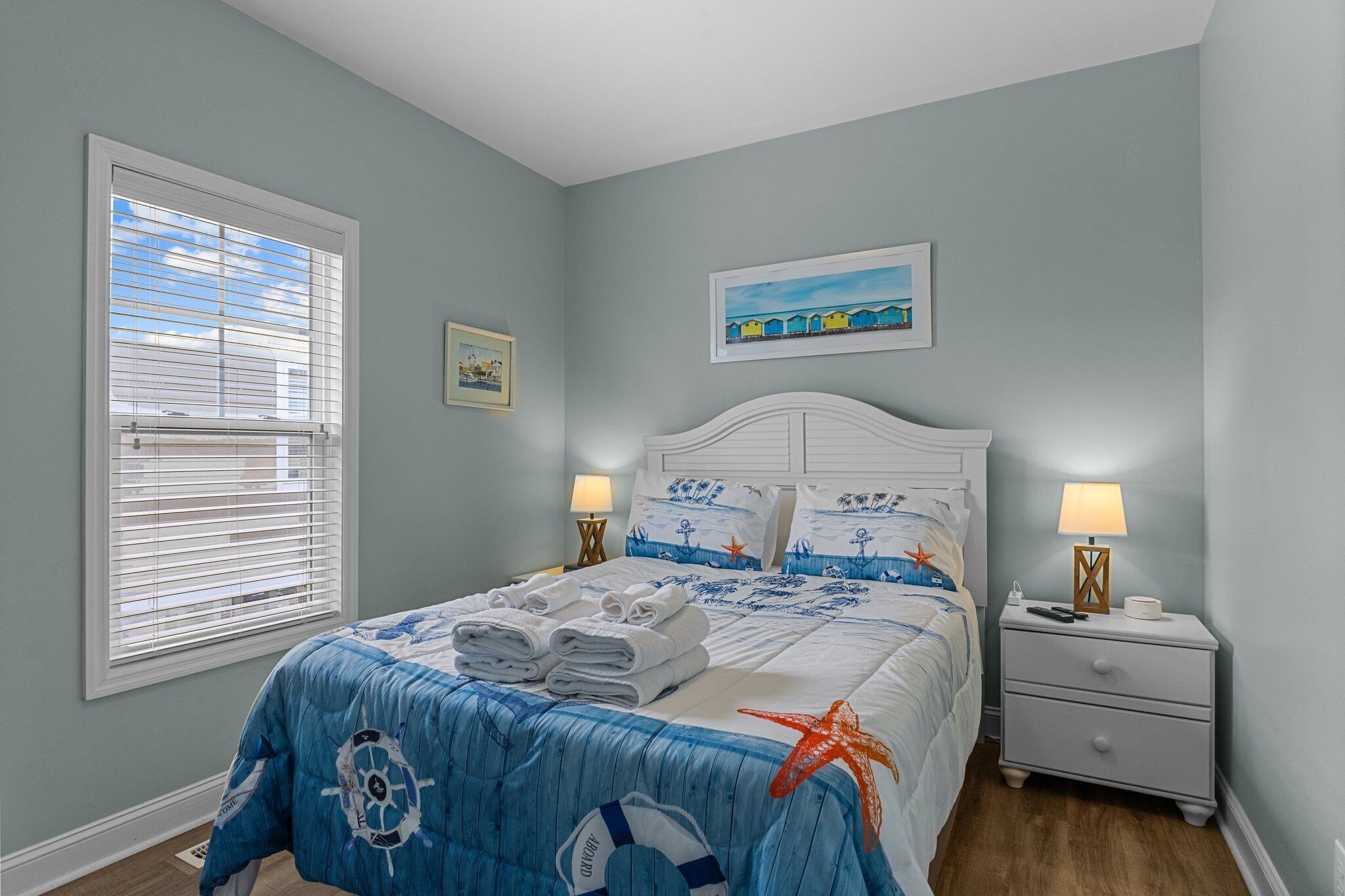 A bedroom with a bed, nightstands, a window with blinds, and a painting on the wall.
