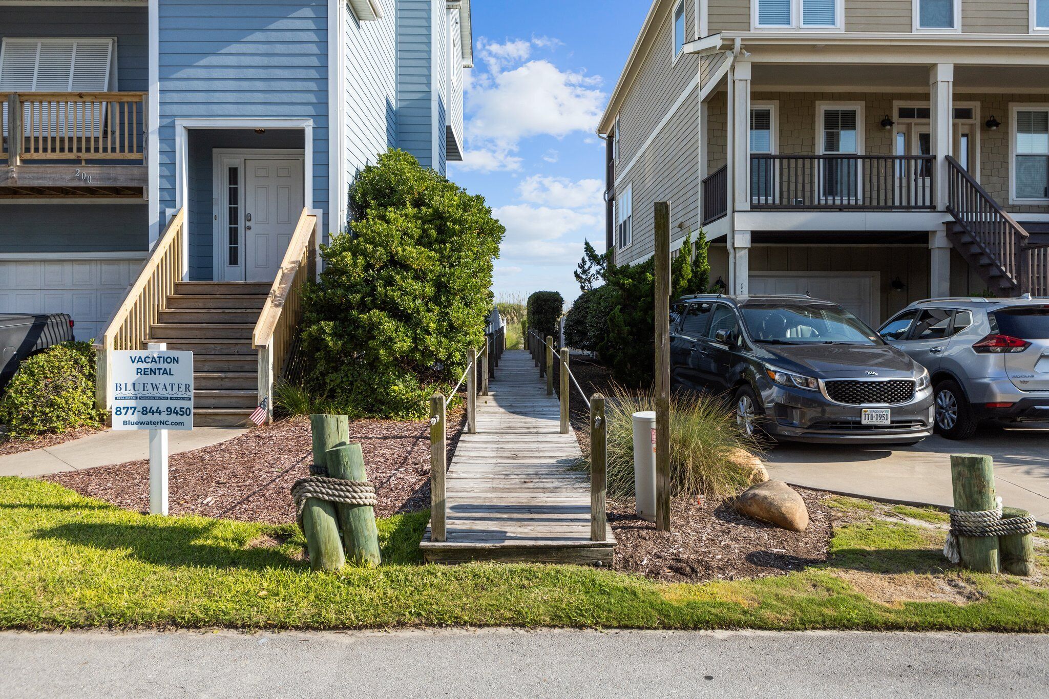 Una calle residencial con un sendero de madera que lleva a una casa. Se ve un cartel de Bluewater Vacation Rental.