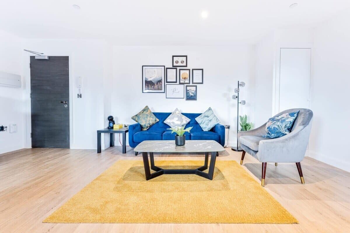 A modern living room with a blue sofa, a grey armchair, a coffee table, and a yellow rug. There are framed pictures on the wall and a door in the background.