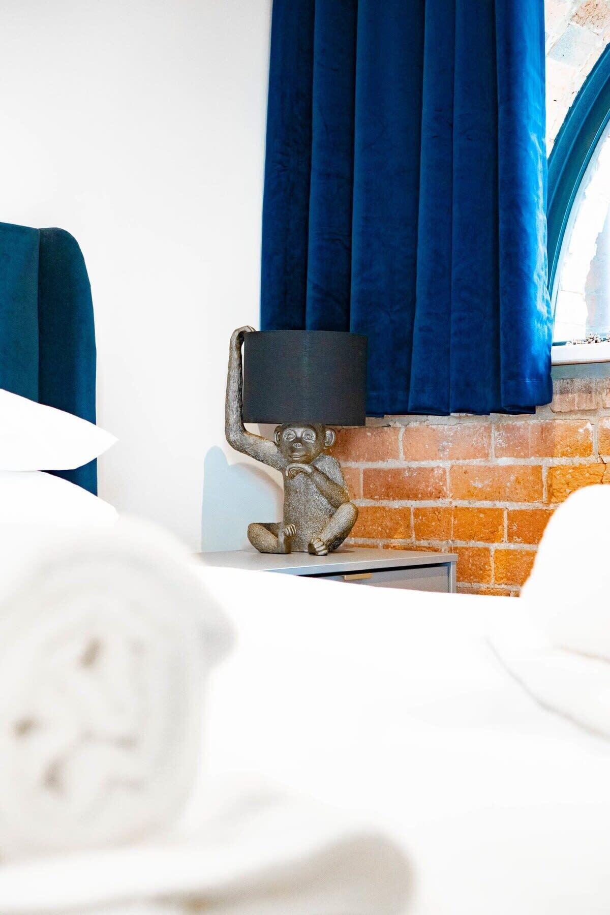 A neatly made bed with white linens, a small table with a lamp, and a brick wall with blue curtains.