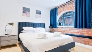 A modern bedroom with a large bed, a nightstand, a lamp, and a brick wall with a window.