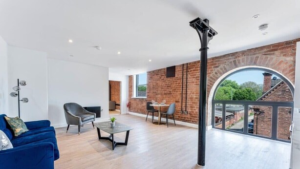 A spacious living room with a blue sofa, a coffee table, and a dining area with chairs. A large window offers a view of a brick wall and greenery outside.