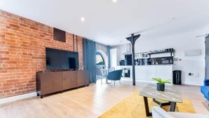 A modern living space with a brick wall, a flat-screen TV, a dining area with a table and chairs, and a kitchen in the background.