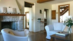 Ein geräumiges Wohnzimmer mit Kamin aus Stein, Holzbalkendecke, weißem Sessel, gemustertem Sofa und Couchtisch mit Buch und Vase.