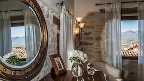 Ein Zimmer mit Steinsturz, einem Holztisch mit Spiegel, einer Vase mit Blumen, einem gerahmten Bild und Blick auf Berge durch das Fenster.