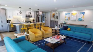 Una sala de estar moderna con un sofá azul, sillas amarillas y una cocina al fondo.