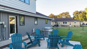 Una casa con un patio que cuenta con cuatro sillas Adirondack azules, una parrilla y una puerta que da a un garaje.
