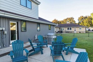 A house with a patio area featuring four blue Adirondack chairs, a grill, and a door leading to a garage.