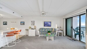 Een ruime woonkamer met een grote schuifpui die leidt naar een balkon, een witte bank met bloemenprint en een kleine bijzettafel met een lamp.