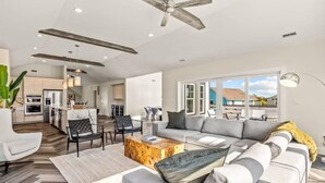 A modern living room with a sofa, coffee table, and chairs. There is a kitchen in the background with cabinets and appliances. - Aurora Elevator Theater Rec Room, Pool & Hot Tub (Corolla)