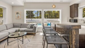 A modern living room with a gray sectional sofa, a glass coffee table, and a kitchen area with a bar counter and a microwave. - Aurora Elevator Theater Rec Room, Pool & Hot Tub (Corolla)