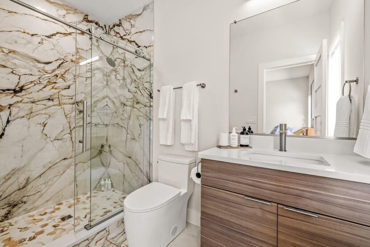 Un bagno con parete doccia in marmo, un water bianco, un mobile lavabo in legno con specchio e un portasciugamani.