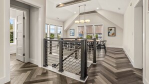 A spacious interior with a herringbone wood floor, a dining area with a table and chairs, and a staircase with a railing. - Aurora Elevator Theater Rec Room, Pool & Hot Tub (Corolla)