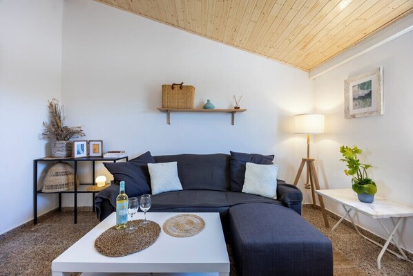 A cozy living room with a dark grey sofa, a white coffee table with a bottle and glasses, a small shelf with decorative items, and a lamp on a side table.