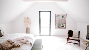 A spacious, well-lit bedroom with a large bed, a bench, a chair, and a door leading to another room. - Sycamore Hill Guesthouse (Oklahoma County)