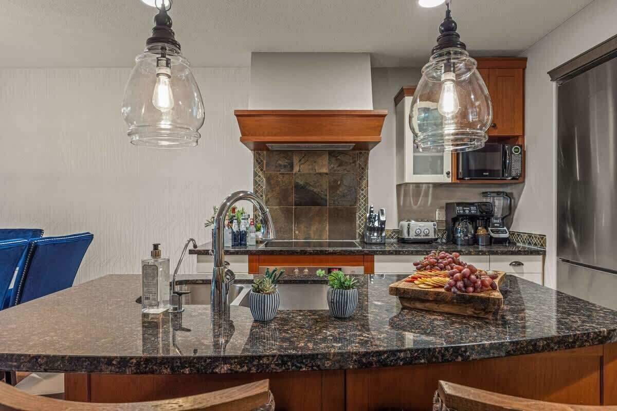 Une cuisine moderne dotée d’un îlot central, d’un plan de travail en granit, d’appareils électroménagers en acier inoxydable et de luminaires suspendus.