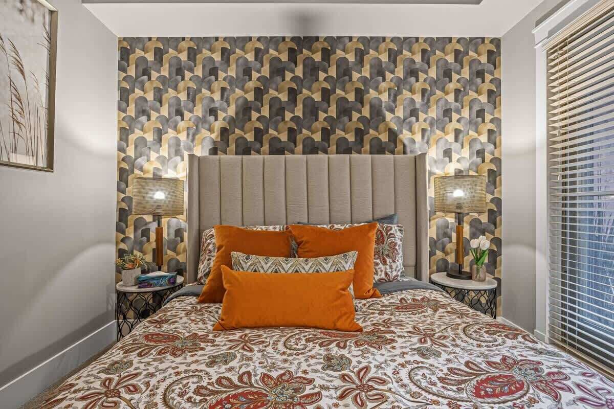 A bedroom with a patterned wallpaper, a bed with orange and patterned bedding, two bedside tables with lamps, and a window with blinds.