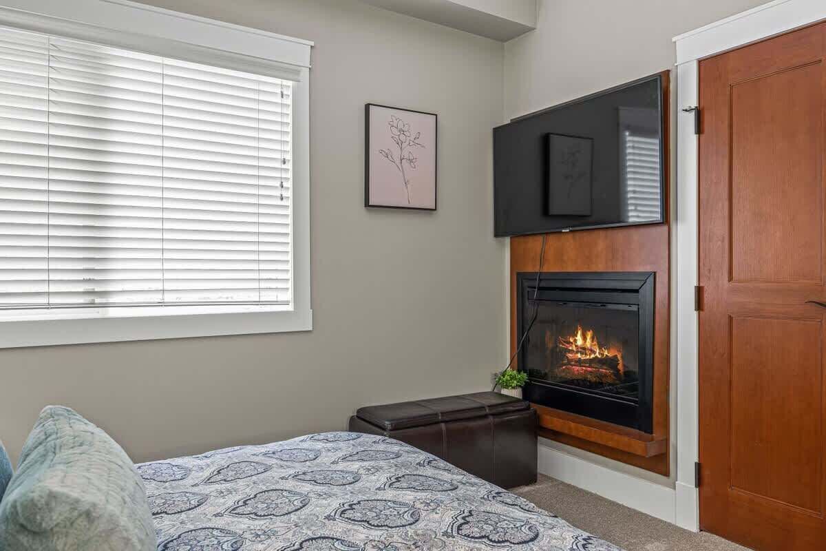 A bedroom with a bed, a fireplace, a TV mounted on the wall, and a window with blinds.