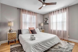 A modern bedroom with a large bed, two bedside tables, a ceiling fan, and sheer curtains.