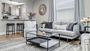 A modern living room with a grey sofa, a glass coffee table, and a kitchen area with a stove and bar stools.