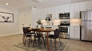 Cocina moderna con armarios blancos, electrodomésticos de acero inoxidable y una zona de comedor con mesa y sillas de madera.