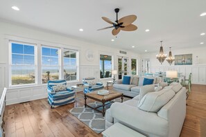 Un vaste salon agrémenté d’un grand ventilateur de plafond, un coin repas illuminé par un lustre, et une vue sur la plage grâce à de grandes fenêtres.