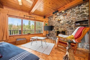A cozy cabin interior with a stone fireplace, wooden beams, a TV mounted on the wall, and a balcony with a view of trees. - Chico | Riverside | Massive Backyard | Fire Pit | (BANCROFT)
