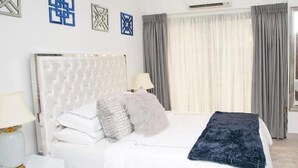 Una camera da letto moderna con una testiera trapuntata, un letto bianco, cuscini grigio e bianchi, una coperta in tessuto blu scuro e un comodino con una lampada.