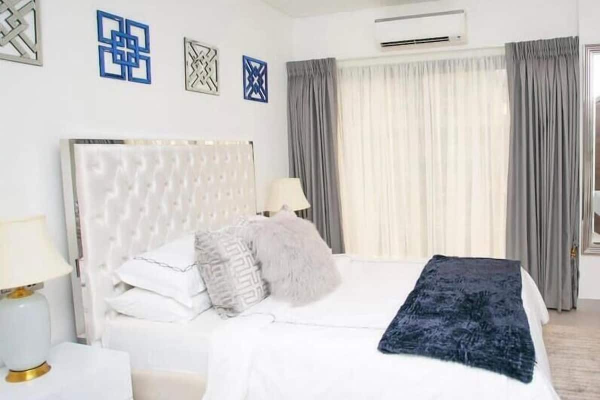 Un dormitorio moderno con cabecera capitonada, cama blanca, almohadas grises y blancas, manta de color azul oscuro y una mesita de noche con lámpara.