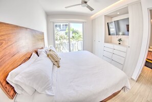 A bedroom with a large bed, a ceiling fan, a TV mounted on the wall, and a sliding door leading to a balcony. - Palmas 101: 1BR w Rooftop Pool, 1 Block to Beach (Bucerías)