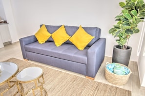 A grey sofa with three yellow cushions, a small round table with a gold frame, a woven rug, a potted plant, and a woven basket.