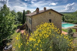 A stone house with a pool, surrounded by greenery and yellow flowers.