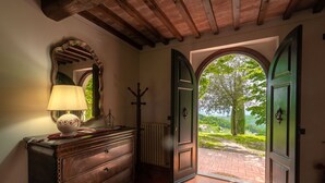 Un couloir avec une commode en bois, une lampe, un porte-manteau et une vue sur un jardin par une porte ouverte.