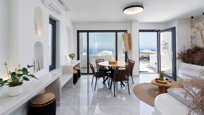 A modern living room with a dining area, large windows, and a view of the sea. - CUORE SANTO SUITES  (Santorini)