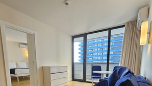 A modern apartment with a large window, a sofa, a small table, and a bed in the background. - iRent Batumi Apartments (Batumi)