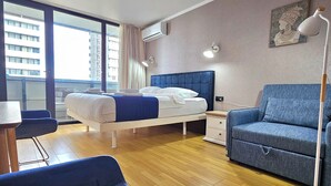 A modern hotel room with a bed, a blue sofa, a desk, and a window with a city view. - iRent Batumi Apartments (Batumi)