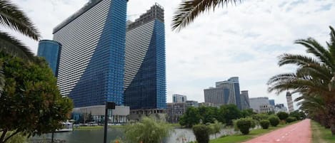 A modern cityscape with high-rise buildings, a calm body of water, and a red pathway lined with greenery.