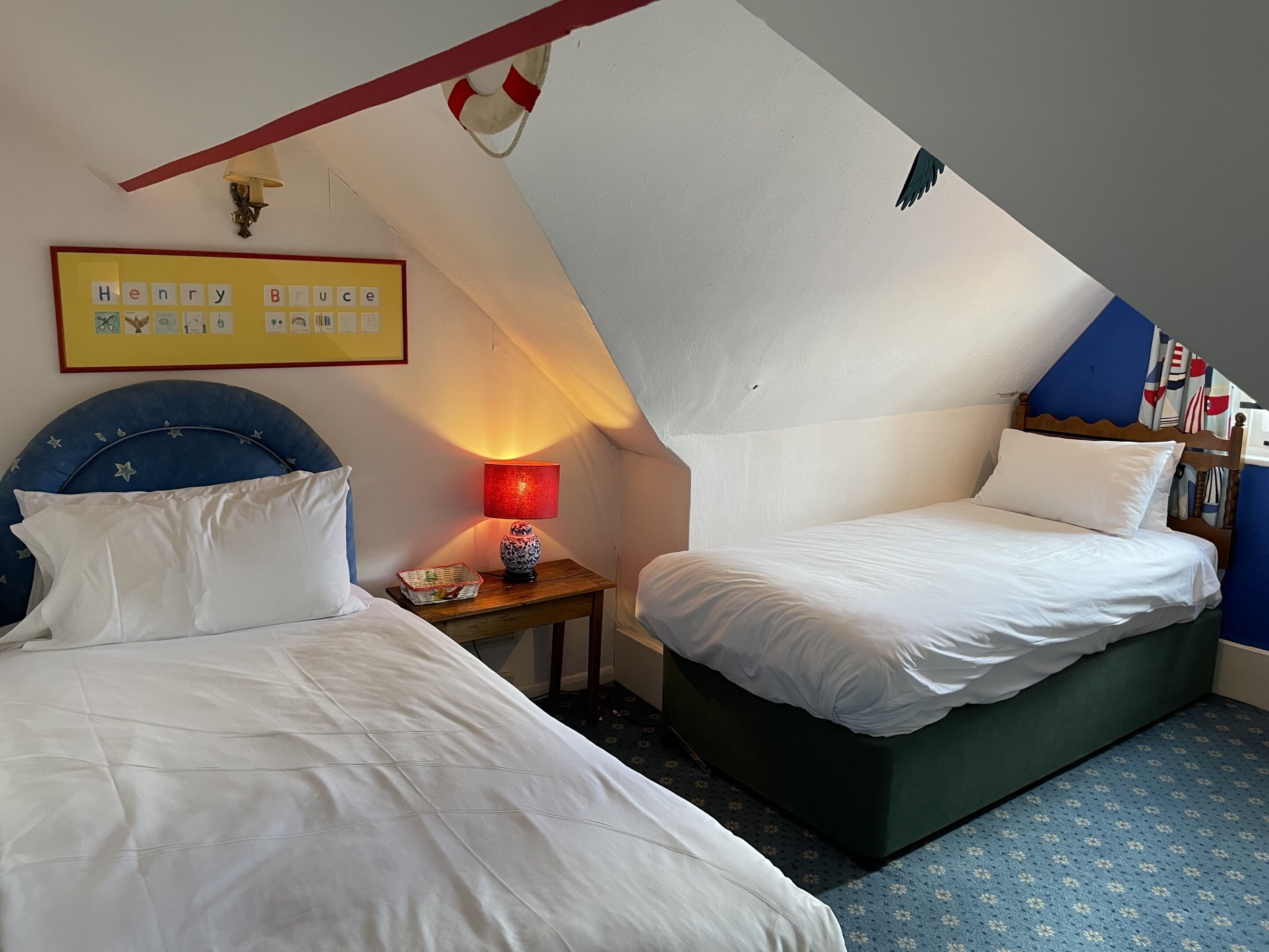 A bedroom with two beds, a nightstand with a lamp, a framed picture, and a ceiling with a red beam and a life preserver decoration.