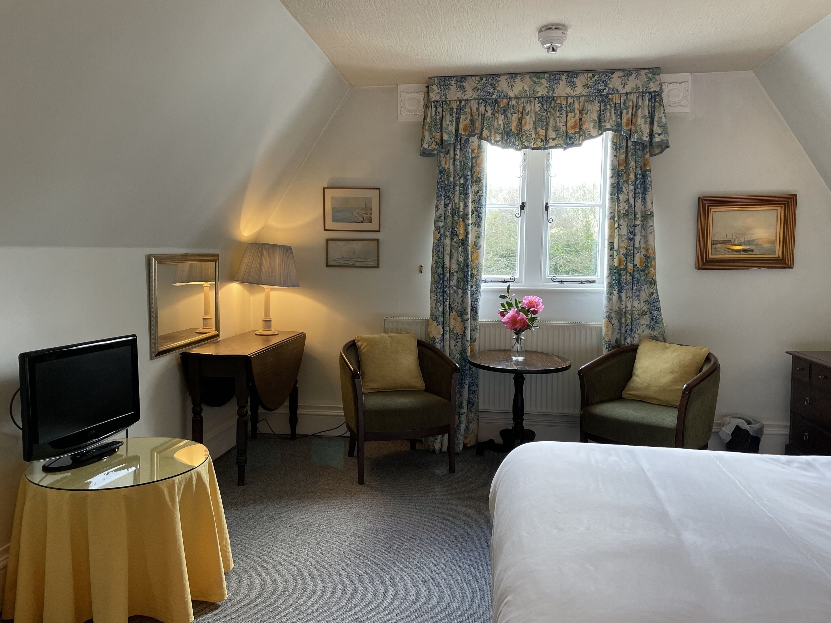 Quarto de hotel com uma cama, televisão, escrivaninha com abajur, duas cadeiras, pequena mesa com um vaso de flores e uma janela com cortinas.