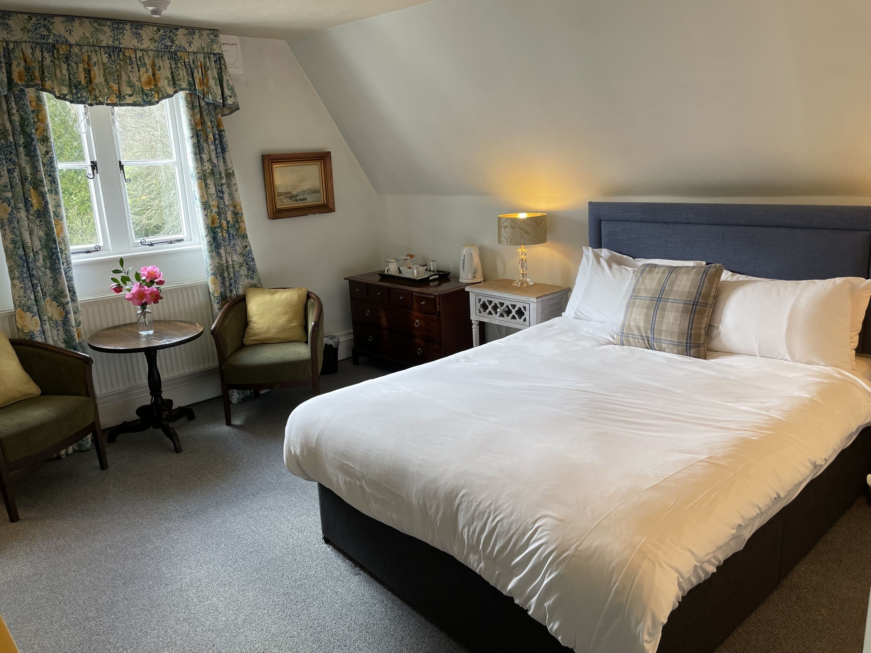 A neatly arranged bedroom with a large bed, a small table with a vase of flowers, a chair, a dresser, and a lamp.