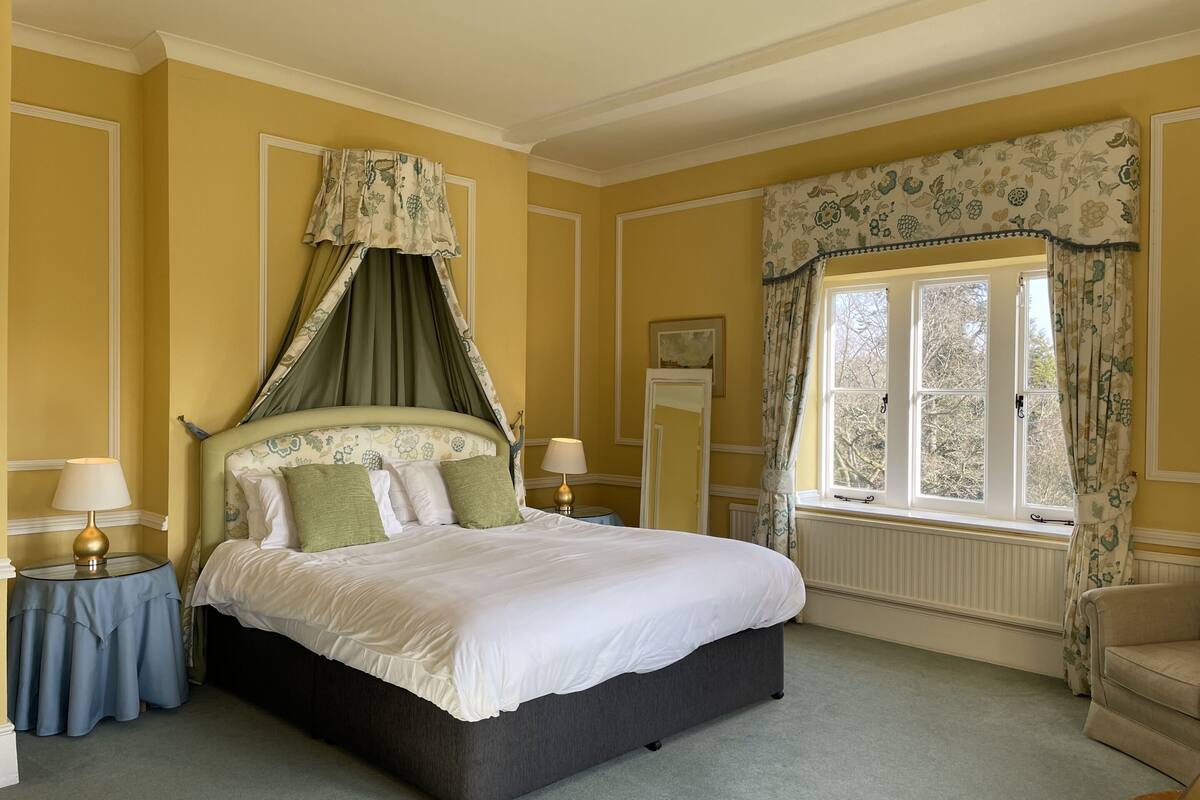 A neatly arranged bedroom with a large bed, a bedside table with a lamp, a chair, and a window with curtains.